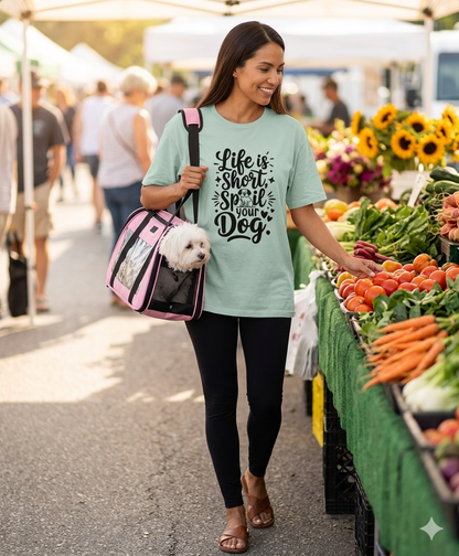 A woman wearing a 'Life is Short, Spoil Your Dog' mint green t-shirt browses a vibrant farmers market while carrying a white Maltese dog in a pink and black travel carrier.